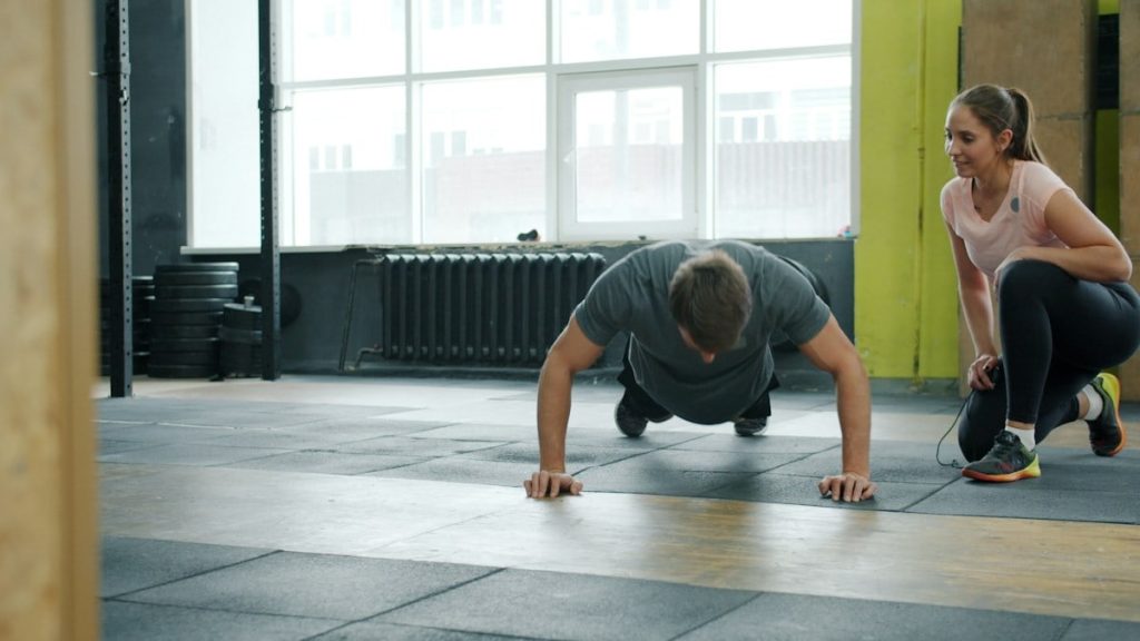 Young man is doing push-ups exercising in gym with female coach enjoying exercise indoors. Active millennials, sports and modern lifestyle concept.