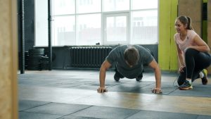 Young man is doing push-ups exercising in gym with female coach enjoying exercise indoors. Active millennials, sports and modern lifestyle concept.